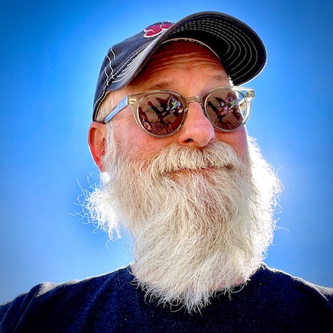 In this photo, Andrew wears a Jeep baseball cap and sunglasses. The sky is bright blue behind him and his long, white beard is backlit by the sun.